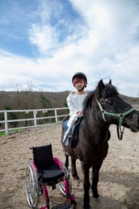 Smiling girl on black pony with wheelchair near